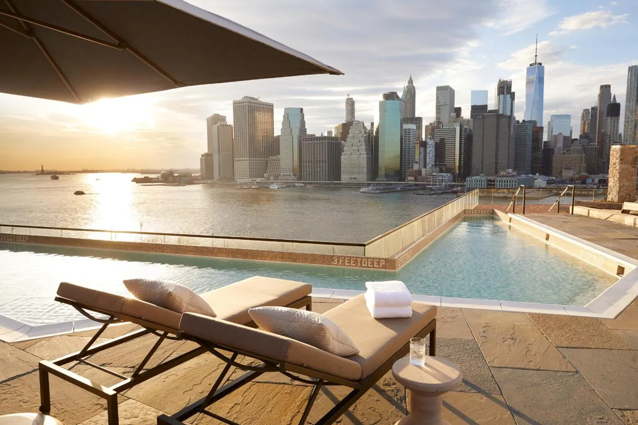 A rooftop infinity pool with sun loungers and a shade umbrella overlooks the Manhattan skyline and river at sunset.