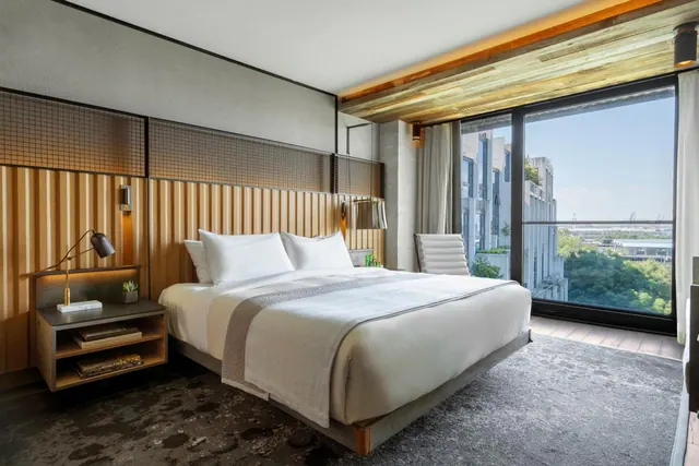 A king bed with white bedding and a bedside table sits beside floor-to-ceiling windows overlooking city buildings and trees.