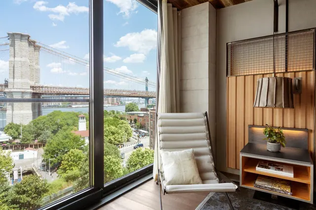 A seating area with a lounge chair and side table beside floor-to-ceiling windows showing the Brooklyn Bridge and the river beyond.