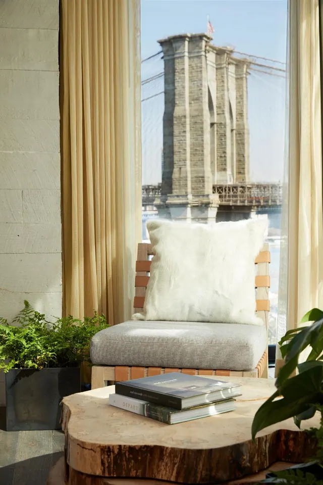 A white fur cushion sits on a grey upholstered lounge chair by a tall window with a view of the Brooklyn Bridge tower, with stacked books on a wooden side table in front.