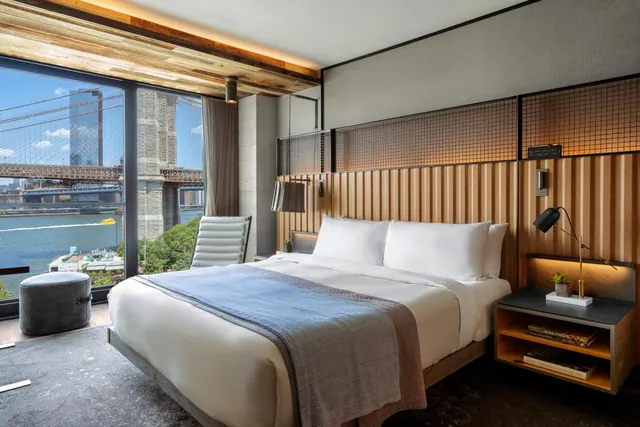 A king bed with white linens faces floor-to-ceiling windows that show the Brooklyn Bridge and the water below.