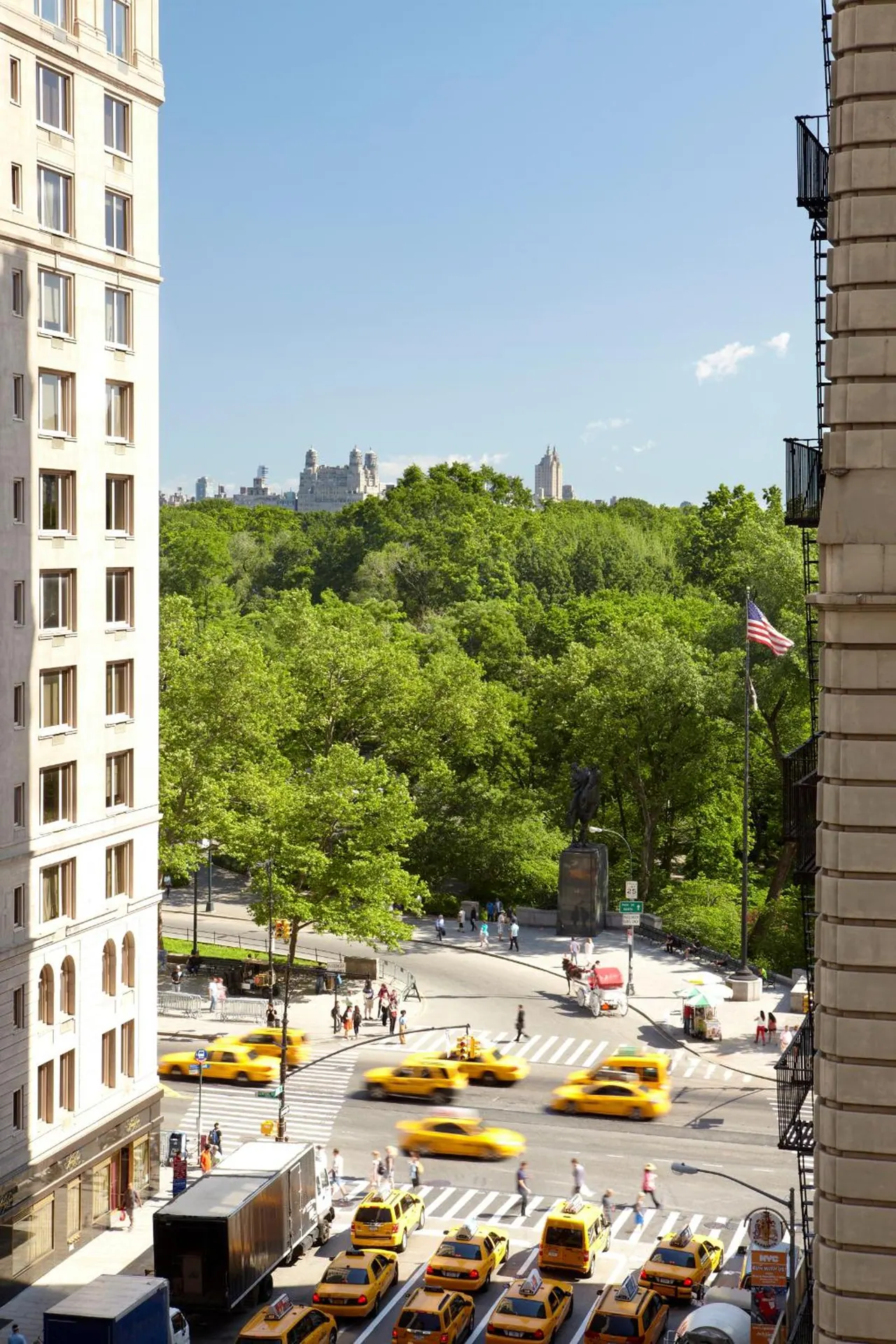 View from an upper floor across a tree-lined park and a street intersection with yellow taxis, toward a bronze statue and distant towers.