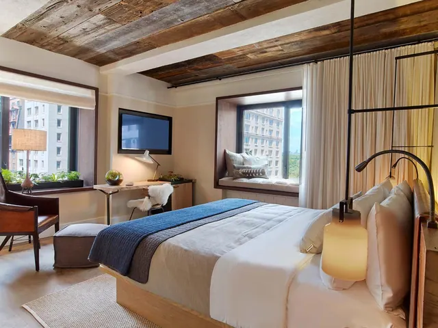 A bedroom with a king bed under a wood plank ceiling, a wall-mounted television, a desk, an armchair and a window seat looking out onto neighbouring buildings.