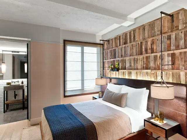 A bedroom with a made king bed, a reclaimed wood accent wall, a window with a roller blind, and a view through the doorway into a bathroom with a vanity.