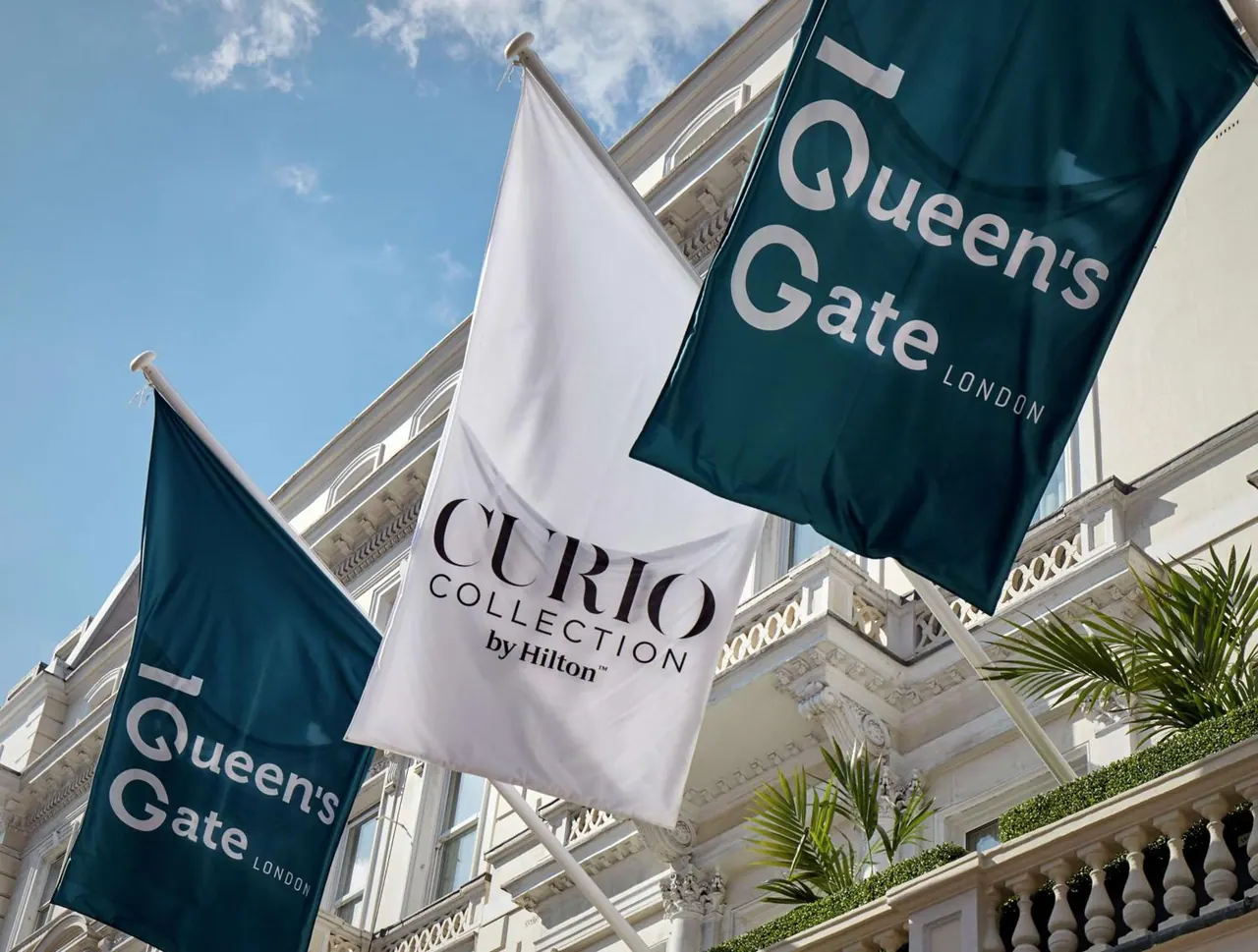 Three flags hang from the white stucco facade of a building, two green ones reading 1 Queen's Gate London and a white one reading Curio Collection by Hilton.
