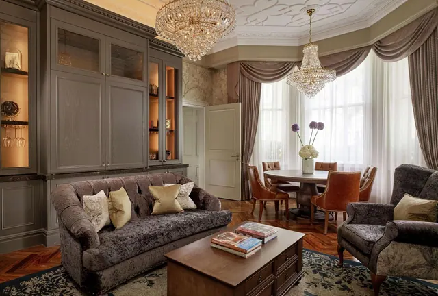 A living area with a tufted sofa, armchairs, a round dining table and two crystal chandeliers beside a tall bay window.
