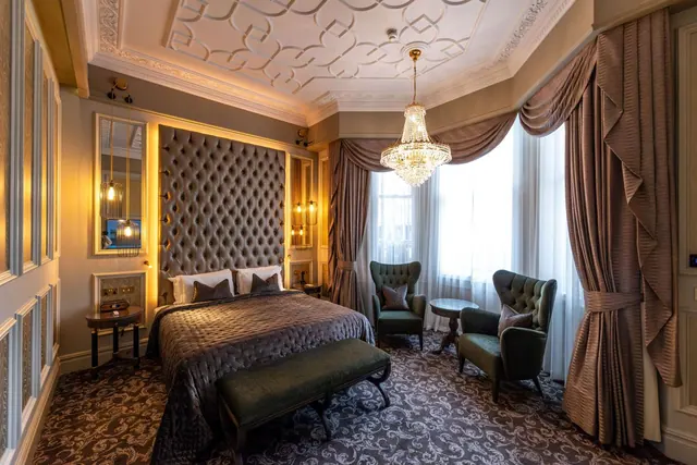 A hotel bedroom with a tufted headboard, a chandelier, two green armchairs by a bay window and an ornate plaster ceiling.