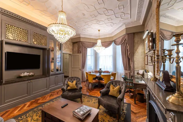 A formal living area with two tufted armchairs, a coffee table, a wall-mounted television in a built-in cabinet, a dining table with yellow chairs, and a bay window with drapes.