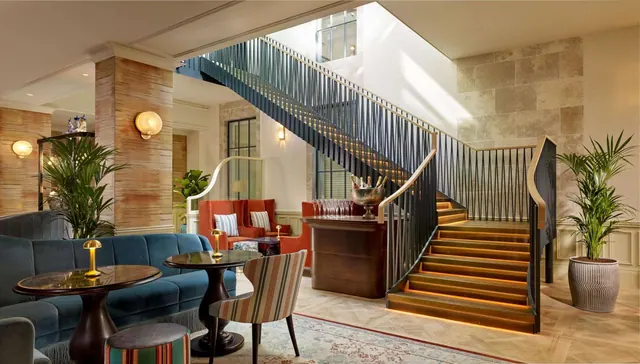 A hotel lobby with a lounge seating area, a bar counter holding a champagne bucket, and a staircase with warm lighting along its treads.