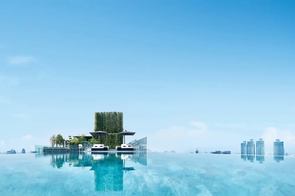 An infinity edge rooftop pool with sun loungers, shade umbrellas and a green wall, with city towers visible on the right.