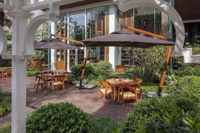 Outdoor dining area on a timber deck with wooden tables, chairs and cantilever umbrellas beside a garden and a glass-fronted building.