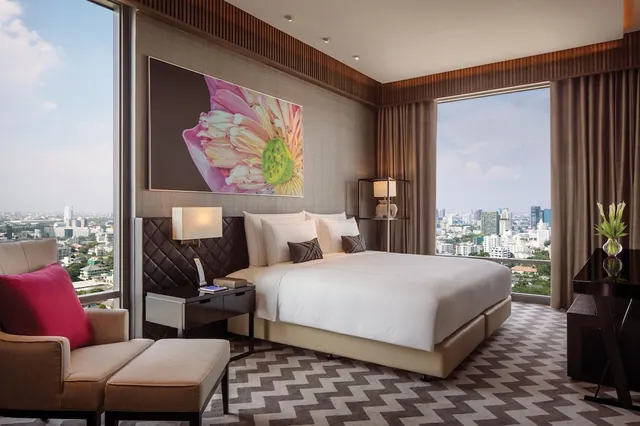 A bedroom with a king bed, an armchair with an ottoman, a desk, and floor-to-ceiling windows looking out over a city skyline.