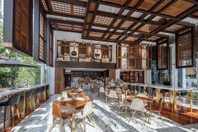 A restaurant dining room with a coffered wooden ceiling, marble floors, wooden tables with white chairs, a bar counter with stools on the left, and greenery visible through the glass wall.