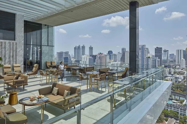 A staff member walks across a high-floor outdoor terrace lounge with brown seating and tables, overlooking a city skyline under a partly cloudy sky.