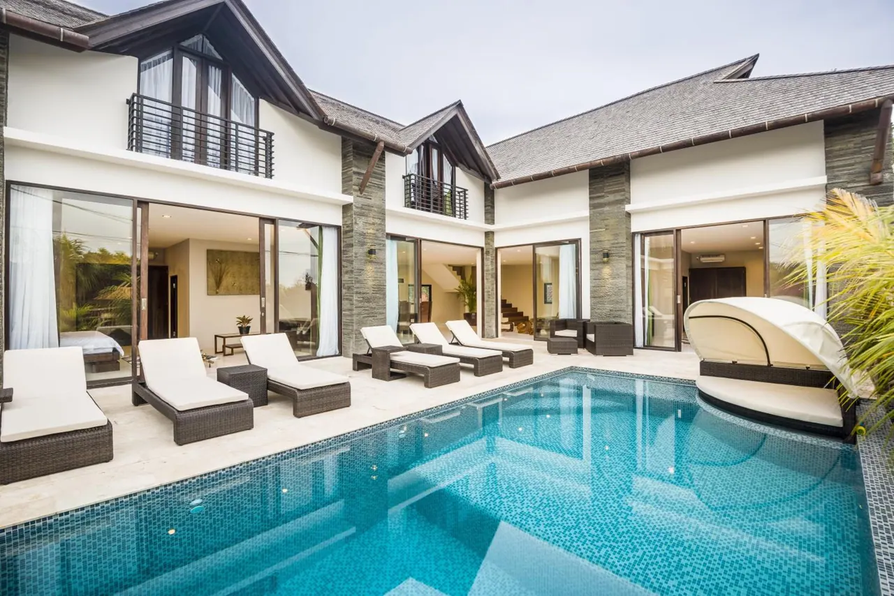 A private swimming pool sits in front of a two-storey villa, with white sun loungers and a canopied daybed on the terrace.