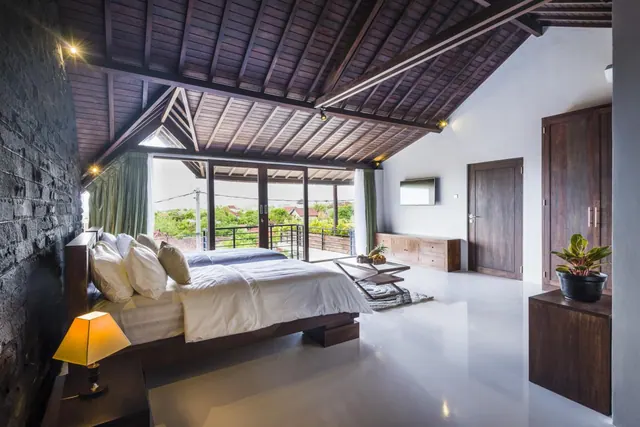 A bedroom with a large bed, an exposed timber ceiling, sliding glass doors opening onto a balcony with greenery beyond, a wall-mounted television and a tall wooden wardrobe.