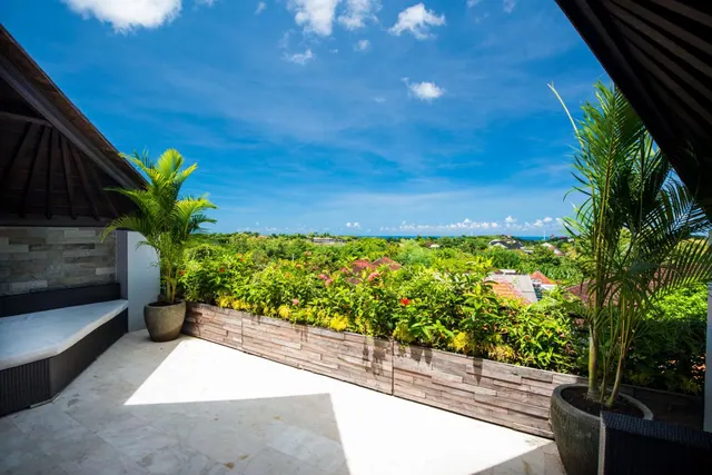 An open-air terrace with stone tiles, a cushioned built-in bench, potted palms and a planter wall, looking over tropical trees and rooftops toward the sea.