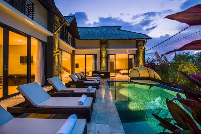 A villa pool at dusk with sun loungers on the terrace and lit rooms visible behind the glass doors.