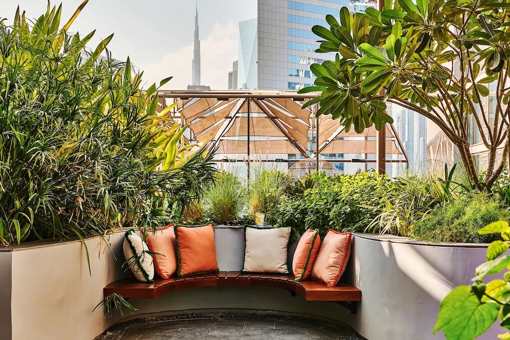 A curved wooden bench with colourful cushions sits among leafy planters on an outdoor terrace, with the Burj Khalifa and other towers visible behind.
