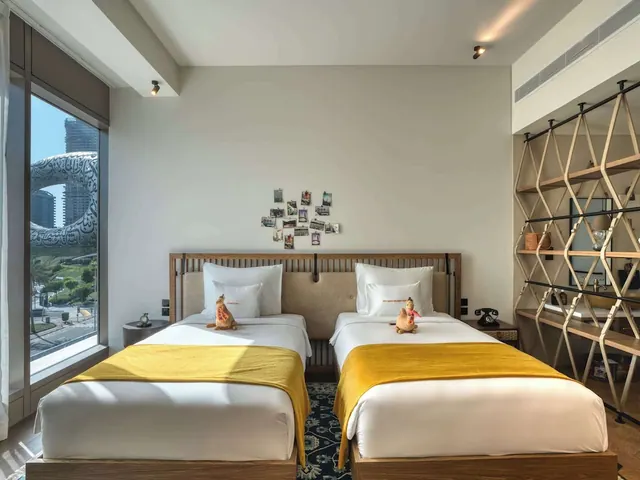 A bedroom with two single beds under yellow throws and plush toys, a tall window showing the Museum of the Future, and open wooden shelving on the right.