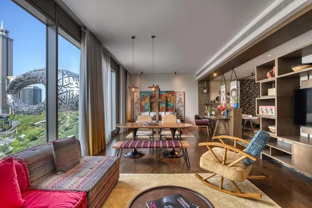 A living area with a patterned sofa, dining table and rocking chair, with floor-to-ceiling windows showing the Museum of the Future and green grounds outside.