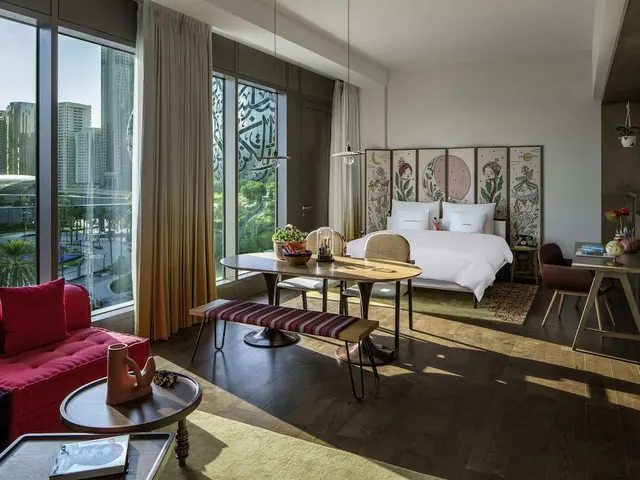 A hotel room with a king bed, a red sofa, a wooden dining table with a bench, and a desk, with floor-to-ceiling windows showing city towers and greenery outside.