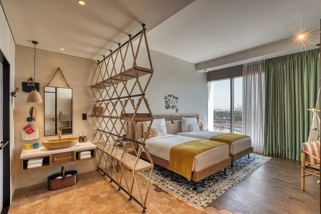 A hotel bedroom with two single beds, a wooden lattice room divider, a vanity basin, and a window looking out over a hazy city skyline.