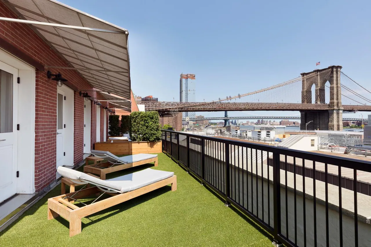 A rooftop terrace with artificial turf and two wooden loungers with white cushions overlooks the Brooklyn Bridge and nearby city buildings.