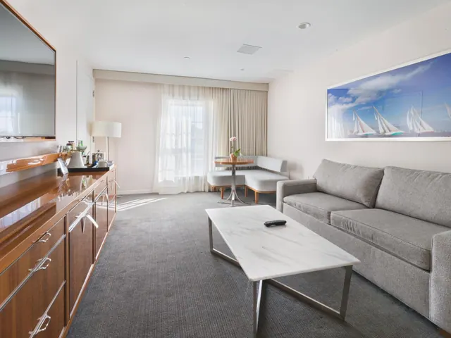 A hotel living area with a grey sofa, a white marble coffee table, a wall-mounted television, a wood sideboard and a framed sailing painting.
