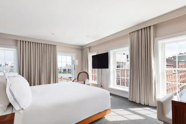 A bedroom with a king bed with white linens, a wall-mounted television, an armchair, a wooden desk, and tall windows looking out over water and city buildings.