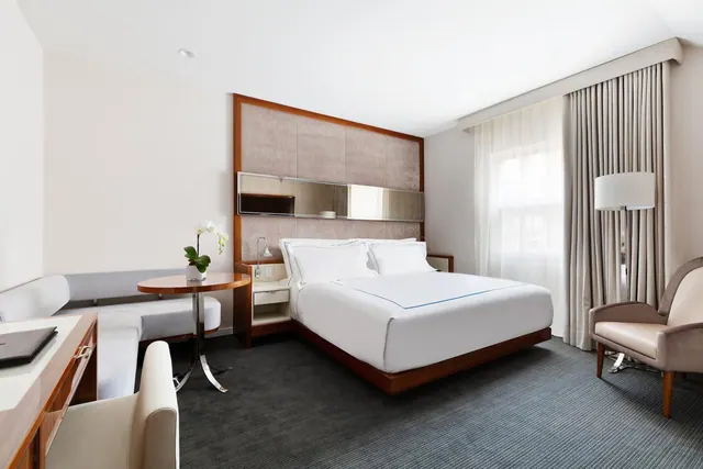 A hotel bedroom with a king bed dressed in white linen, a grey banquette, a round side table holding an orchid, an armchair, and a wooden desk near the entrance.
