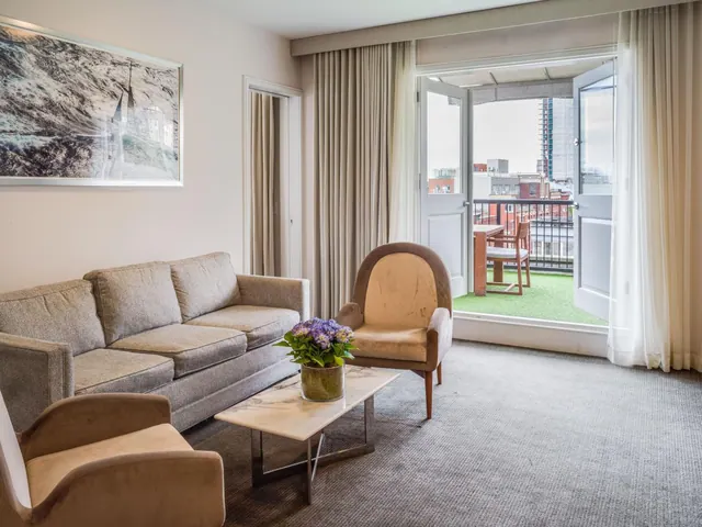 A living area with a grey sofa, two armchairs and a marble coffee table holding a flower arrangement, with open doors onto a balcony overlooking city buildings.