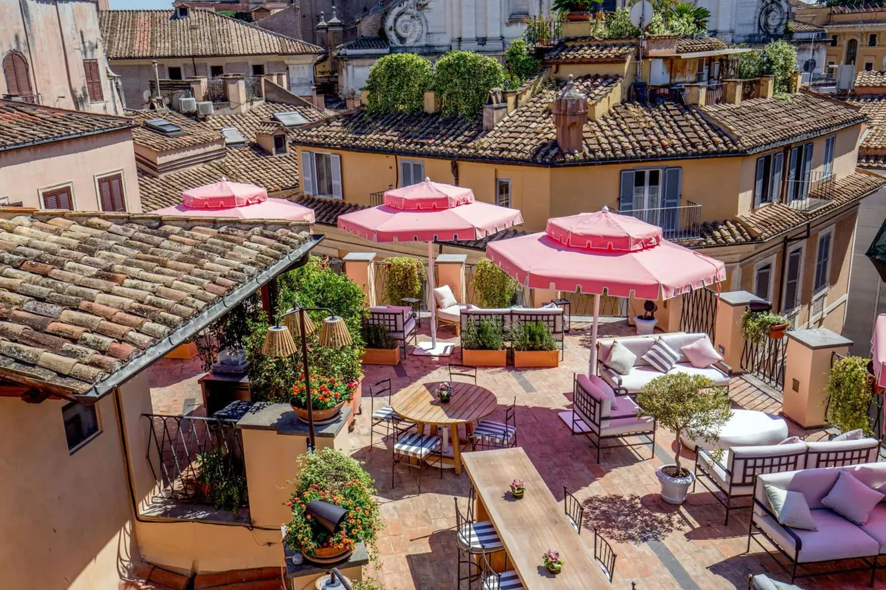 A rooftop terrace with pink umbrellas, sofas and dining tables overlooks terracotta rooftops and a church facade.