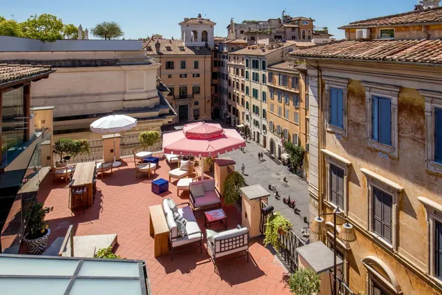 A furnished rooftop terrace with sofas, armchairs and pink and white umbrellas overlooks a street and historic ochre buildings with terracotta roofs.