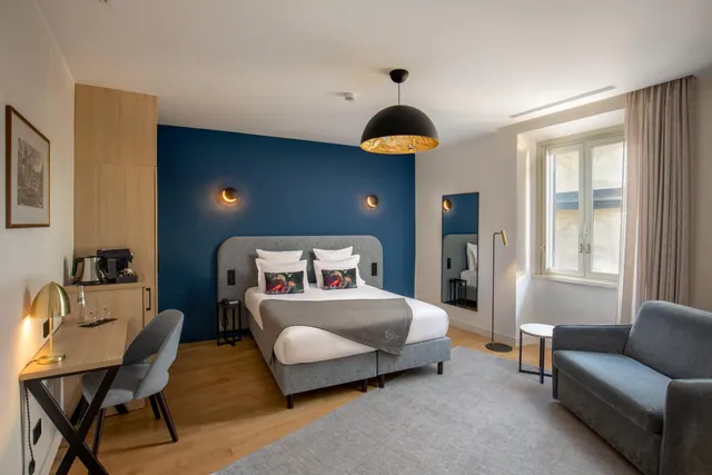A hotel bedroom with a double bed against a deep blue accent wall, a wooden desk with a chair, a grey sofa, and a kettle and coffee machine on a wooden cabinet.
