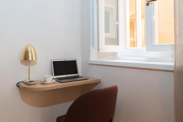 A small wall-mounted desk holds a laptop, a brass lamp and a cup on a saucer, beside a window with a chair pulled up to it.