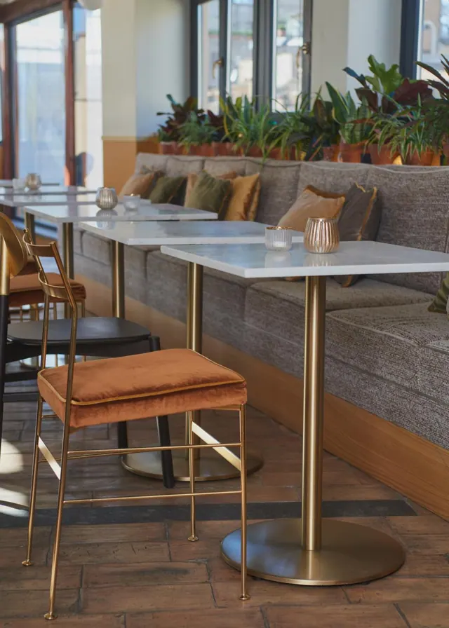 Empty dining area with marble-topped tables on gold pedestal bases, a grey upholstered banquette with cushions, potted plants behind it, and orange velvet stools in the foreground.
