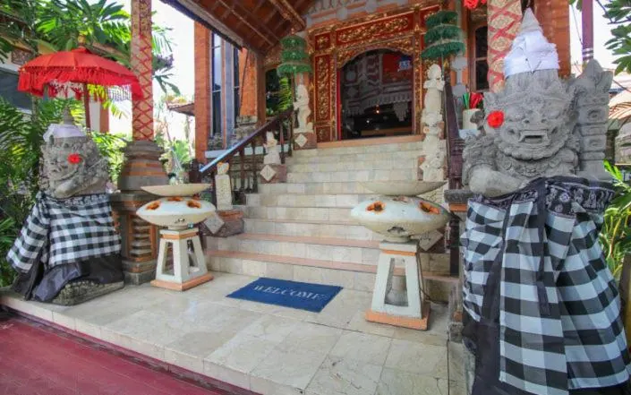 Entrance of a hotel with two stone guardian statues in checkered sarongs, flower bowls on pedestals, and steps leading to a carved red and gold doorway.