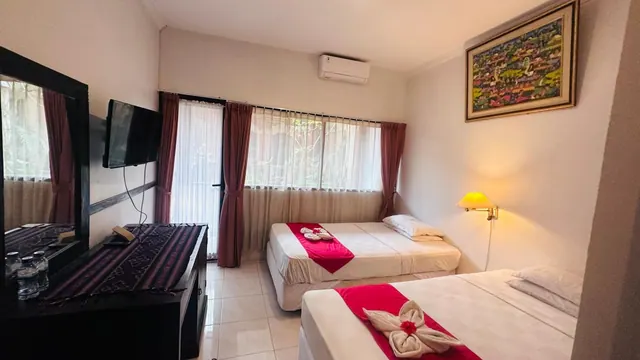 A hotel bedroom with two beds dressed with red runners and towel swans, a wall-mounted TV, an air conditioner and a framed painting.