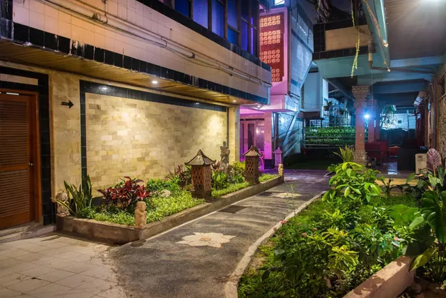 A night view of a paved walkway lined with planted beds, stone lanterns and statues, with hotel buildings on either side.