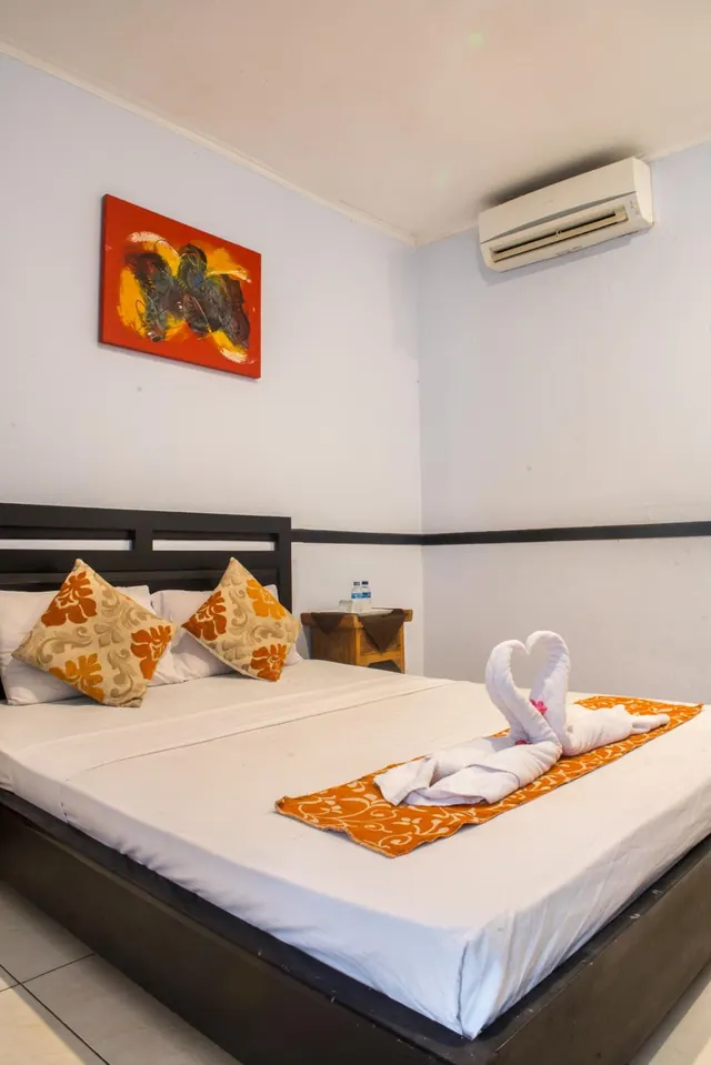 A double bed with orange patterned cushions and a folded towel swan on an orange runner sits below a wall-mounted air conditioner.