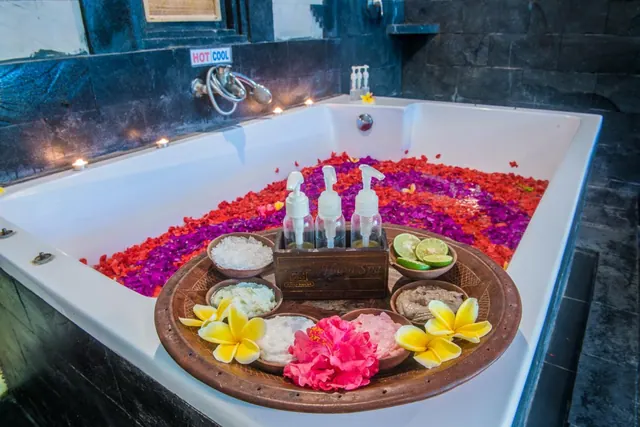 A bathtub filled with red and purple flower petals, with a wooden tray of spa products, pump bottles, bowls of salt and scrub, and frangipani flowers in front.