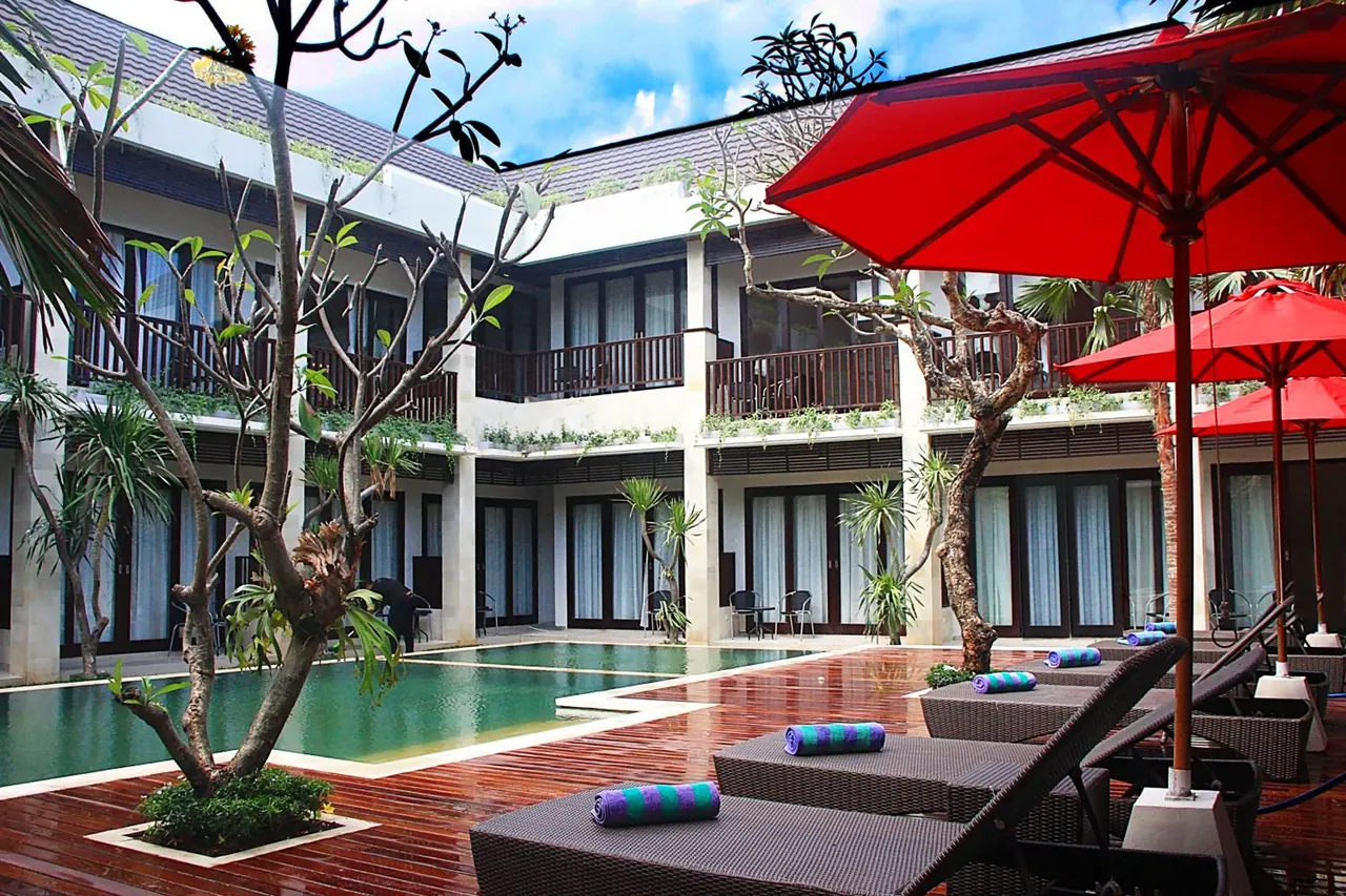 A courtyard pool with red umbrellas and sun loungers on a timber deck, with two-storey rooms with glass doors behind.
