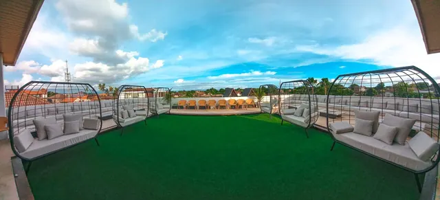 A rooftop terrace with green artificial turf, caged egg-shaped lounge seats with cushioned sofas, and rows of orange chairs on a raised deck under a partly cloudy sky.