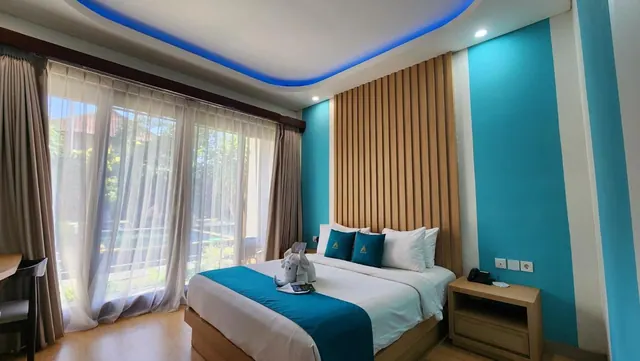 A bedroom with a made king bed, teal accent wall, wooden slatted headboard, sheer curtains and glass doors looking onto greenery.