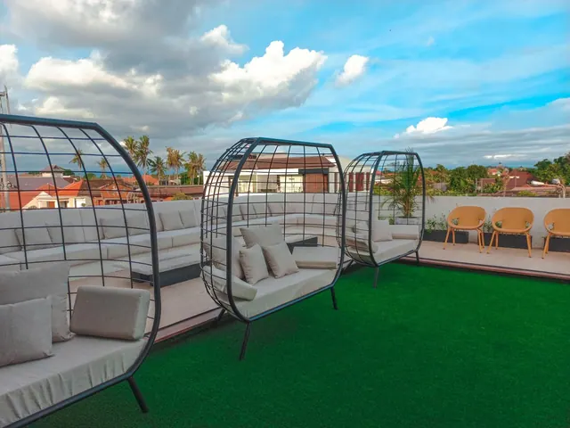 A rooftop terrace with green artificial turf, caged outdoor lounge seats with cushions, orange chairs and planters, with rooftops and palm trees visible beyond the wall.