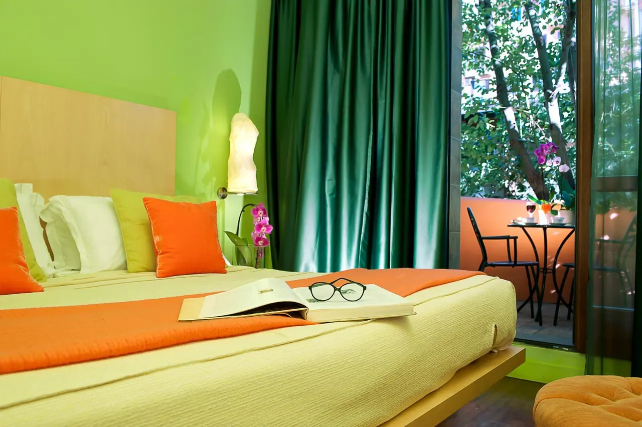 A bedroom with green walls and a bed with orange and yellow bedding, an open book and glasses on the bed, and a glass door opening onto a balcony with a table and chairs among trees.