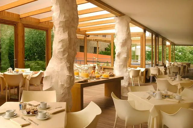 A breakfast dining room with white sculptural columns, set tables, a central table with fruit and juice, and glass walls looking onto green trees.