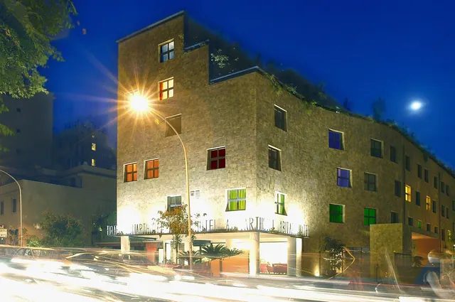 Night exterior of a stone-faced hotel building with coloured windows, plants along the roofline, a street lamp, and blurred traffic in the foreground.
