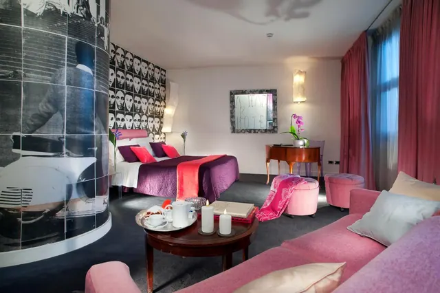 A hotel room with a double bed under purple bedding, a pink sofa and ottomans, a round wooden coffee table holding a tray and two candles, and a large black-and-white photo panel on the left.